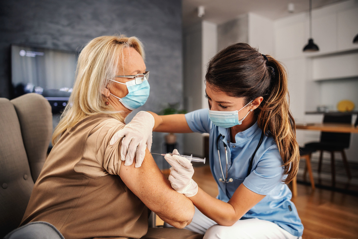 Nurse with protective face mask on sitting at home and giving injection to an old woman during corona outbreak. Nurse performing an at home vaccination
