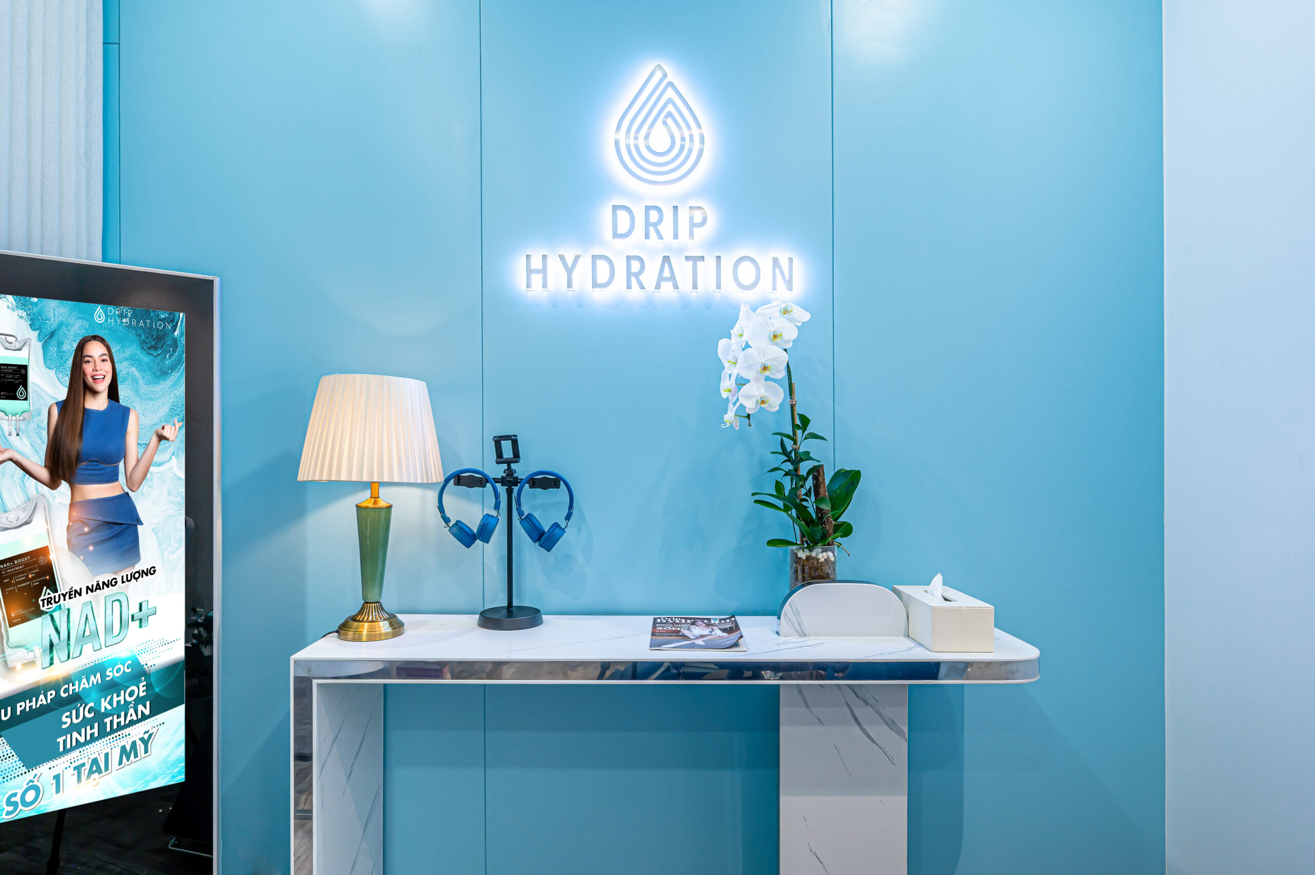 neon sign that says "Drip Hydration" over a desk with a lamp, headphones, plants and a tissue container neon sign that says "Drip Hydration" over a desk with a lamp, headphones, plants and a tissue container